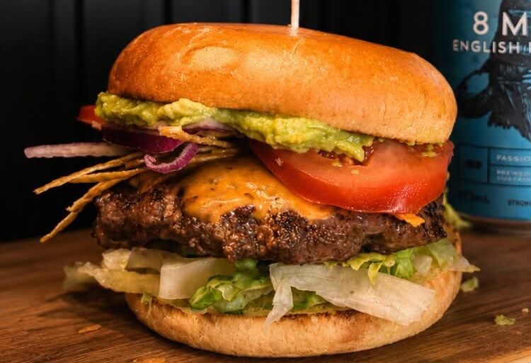The Cantina Burger with avocado, tomato and crispy onions beside an 8 Man pale ale can on a dark wall
