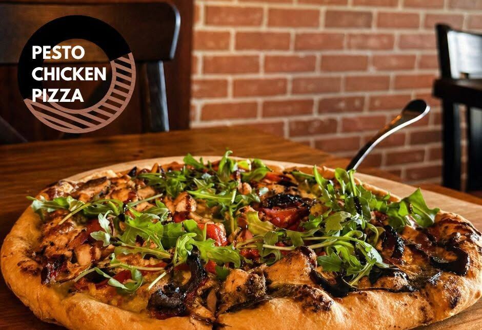 Pesto Chicken Pizza topped with arugula, a whole pie on a wooden table with a brick wall behind