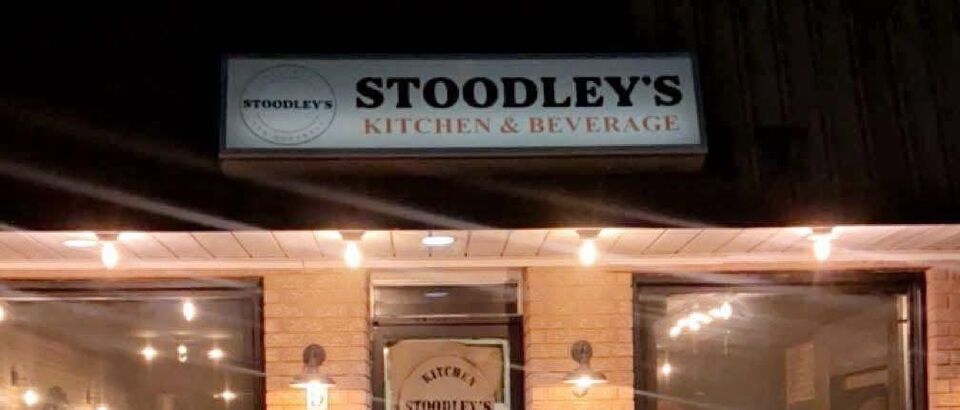 The front of Stoodley’s at night: the lit sign reading Stoodley’s Kitchen and Beverage, string lights along the eave, brick and warm windows